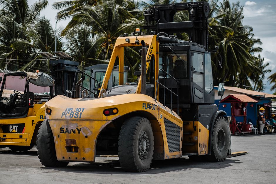 A yellow SANY forklift loaded with large wooden pallets used for packing and moving, positioned on a paved area in an outdoor setting with several palm trees and partly cloudy sky in the background. The forklift has a protective cab with glass windows and black accents, and its forks are lowered to the ground, ready for transport. Nearby, there is another construction vehicle, painted yellow as well, with visible branding, and a red and blue building with a few workers and other machinery in the background. This scene depicts equipment used in the logistics of home relocation and furniture transport, emphasizing the heavy machinery involved in loading and unloading goods during a moving process. The overall environment suggests an outdoor equipment yard or staging area, with elements relevant to the logistics of packing, loading, and transporting household items, aligning with the services offered by Man with Van Teddington for residents in the TW11 area, especially in regards to parking and loading bay solutions for moves.