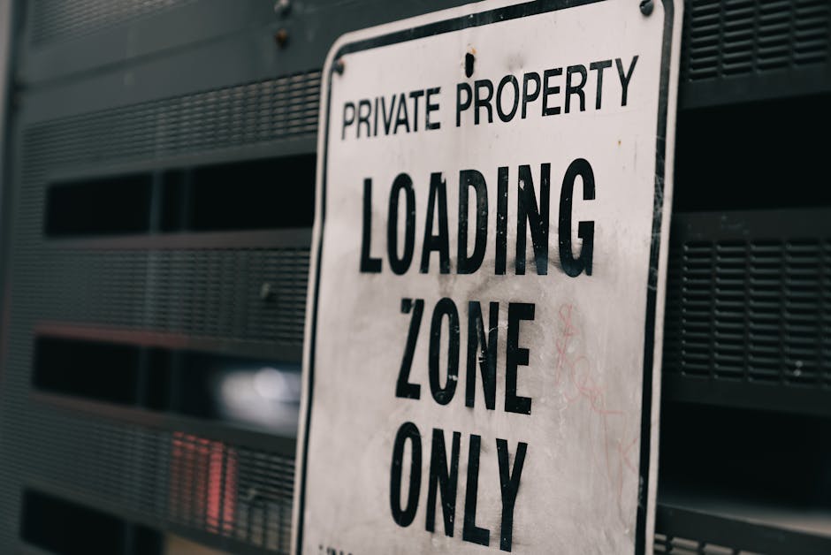 Close-up photograph of a white rectangular sign with black text reading 'PRIVATE PROPERTY LOADING ZONE ONLY', mounted on a dark grey metal wall with horizontal slats. The sign is positioned slightly tilted to the right, indicating a restricted area designated for loading activities during home relocation or furniture transport. The background shows the metallic surface of the wall, which appears weathered and slightly rusted, suggesting an outdoor setting near a property entrance. This image reflects the importance of complying with parking and loading regulations in residential moving processes, often managed by professional removals companies like Man with Van Teddington, especially when navigating tight urban spaces or restricted zones.