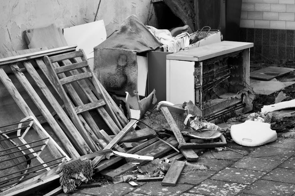 A collection of discarded household items and debris piled outside a property on a paved surface. The pile includes wooden ladders, broken pieces of furniture, a damaged white cabinet with its doors removed, and various detached panels and boards. There are also plastic and cardboard packaging materials, along with a white plastic item that appears to be a removed toilet seat or similar object. The objects are arranged haphazardly, partially leaning against a wall and scattered across the pavement, indicating a cleanup or removal process related to house clearance or renovation. This scene appears to be part of a home relocation or furniture transport project handled by Man with Van Teddington, illustrating the clearance of bulky waste prior to moving or disposal.