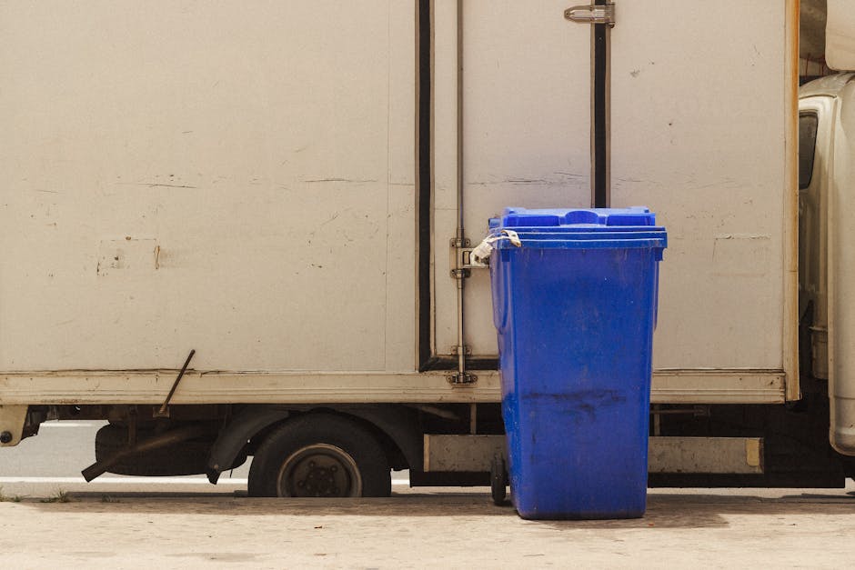 A section of a white delivery van parked on a paved surface, with the side door partly visible. In front of the van, a large blue recycling bin with a closed lid is positioned on the ground, close to the van's sliding door. The van's interior is not visible, but the edges of the vehicle show signs of wear and dirt. The scene appears to be part of a house relocation or moving process, where the removal of bulky waste or recyclable materials, such as the blue bin, is underway. The lighting is neutral, indicating daytime, with no people present in the image. The setting underscores the logistics involved in packing up and moving household contents, with a focus on handling large or bulky waste items as part of a broader move, as managed by Man with Van Teddington, operating within the realm of removals and home relocation services.