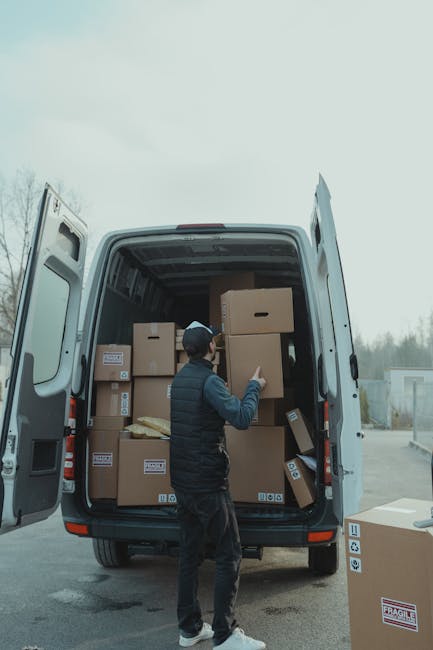 A man wearing a dark jacket, dark trousers, and a backward baseball cap is standing beside an open moving van, which is parked outside on a paved area. Inside the van, numerous cardboard boxes of various sizes are neatly stacked, some wrapped with plastic or packing tape. The man is holding a medium-sized box with both hands, preparing to load or unload it, while other boxes are positioned near the van's interior walls. The background shows a cloudy sky, a few trees, and a distant building, indicating an outdoor home relocation process. Several boxes are situated on the ground nearby, suggesting they are part of an ongoing packing or unpacking activity. The scene captures the logistics involved in furniture transport and packing for a professional house removal service, such as offered by Man with Van Teddington.