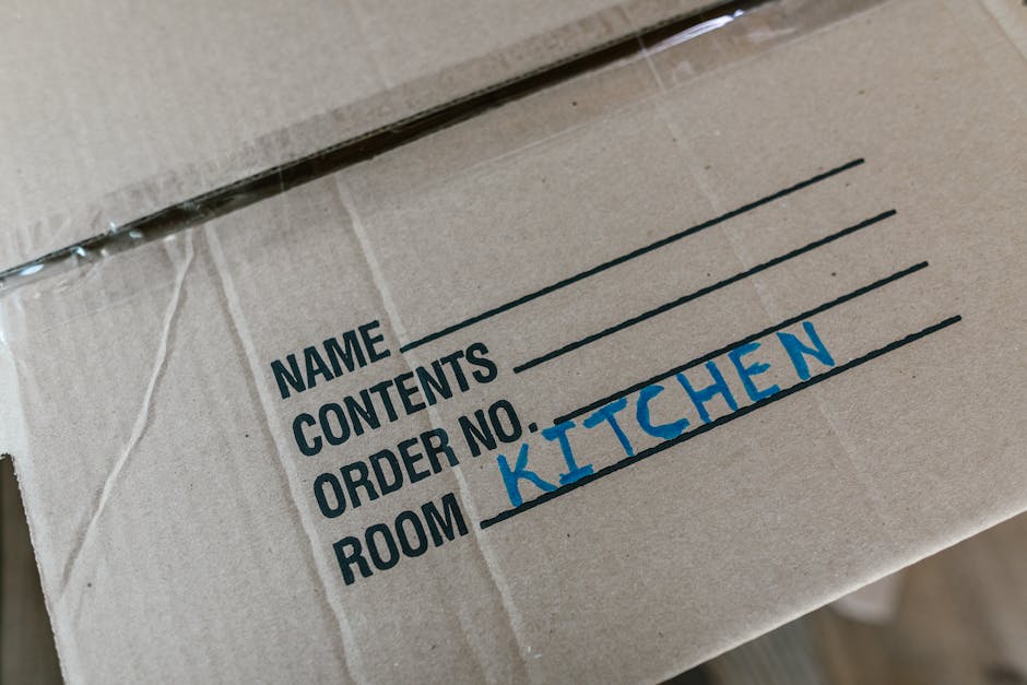 Close-up view of a cardboard moving box with printed labels, including sections for Name, Contents, Order No., and Room, with the room label handwritten in blue ink as 'KITCHEN'. The box appears to be part of a home relocation process, situated on a surface indoors, with other boxes and packing materials possibly visible nearby. The scene suggests the packing stage of a house removal service by Man with Van Teddington, emphasizing careful organisation and preparation for furniture transport and moving logistics.