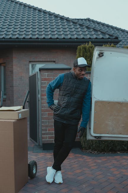 A man wearing a white t-shirt and jeans is seen standing outside on a residential street, loading several cardboard boxes into the rear of a white van parked next to a sidewalk. The van's rear door is