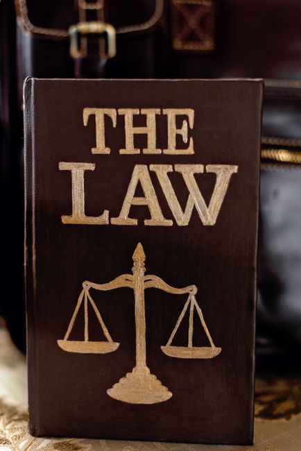 A black hardcover book titled 'The Law' in large gold letters on the front cover, with a gold illustration of balanced scales beneath the title. The book is positioned upright on a surface, surrounded