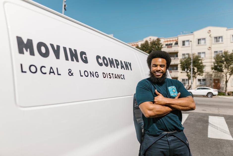 A man with dark skin, a curly afro hairstyle, and a full beard is standing outdoors next to a white moving van with the text 'MOVING COMPANY, LOCAL & LONG DISTANCE' printed on its side. He is wearing a dark teal T-shirt with a logo on the chest, and has his arms crossed while smiling directly at the camera. Behind him, residential buildings with multiple stories and balconies are visible, along with parked cars and trees lining the street. The scene is well-lit with natural daylight, and the image depicts a home relocation or furniture transport process, illustrating a professional removals service provided by Man with Van Teddington, focused on local and long-distance house moves.