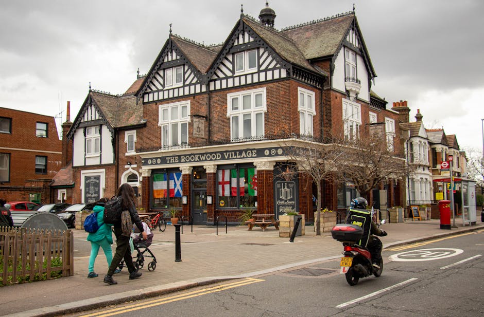 A street scene outside The Rookwood Village pub in Teddington, featuring a two-story brick building with white and black timber framing on the upper floor, and multiple flags displayed above the entrance. In the foreground, a woman pushes a stroller along the pavement, while another person on a black scooter with a top case is waiting or preparing to move. Several parked cars are visible on the side of the street, along with a red post box, a bus stop shelter, and a leafless tree. The surrounding area includes additional brick and white-painted buildings, and cloudy overcast skies above. This scene captures the typical environment of home relocation or furniture transport within a busy suburban neighborhood, with an emphasis on the built environment and pedestrian activity, relevant to house removals and moving services offered by [COMPANY_NAME].