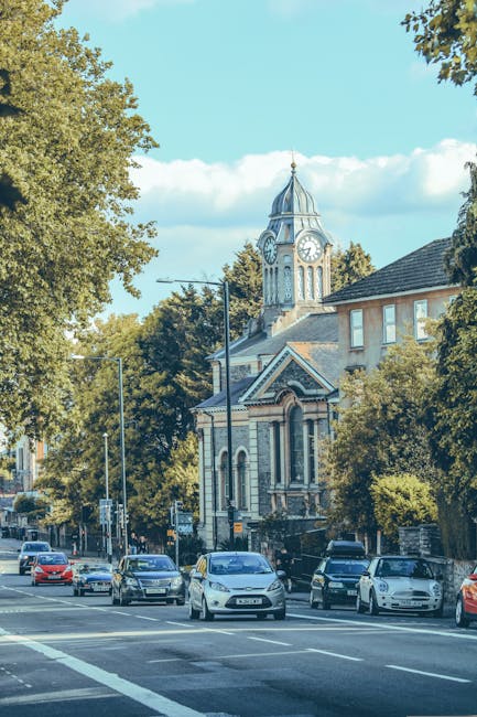 The image shows a street scene in Teddington during daytime with multiple parked cars lining the curb and a few vehicles parked along the road. In the background, there is a prominent historic building with a clock tower, featuring a domed roof and decorative architectural details, surrounded by leafy green trees that partly obscure the building's lower sections. The scene is well-lit with natural sunlight, and the sky is partly cloudy with patches of blue visible. The street appears to be in a residential or central area of Teddington, and this setting is relevant to house removals and moving services offered by Man with Van Teddington, which specializes in home relocation, furniture transport, packing, and loading processes, as depicted in the context of Richmond Council permits for house moves in Teddington.