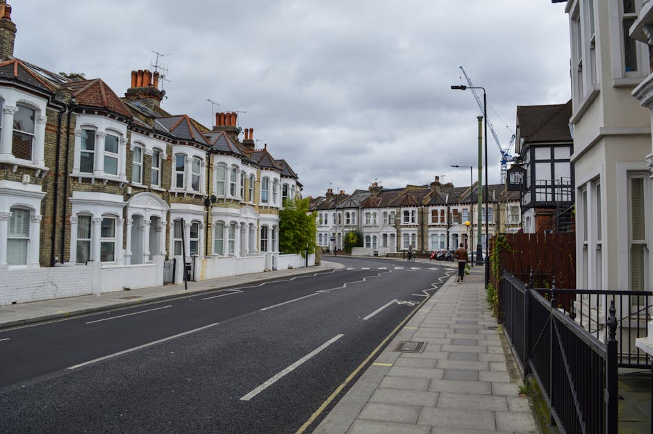 A quiet residential street with a row of Victorian-style terraced houses featuring bay windows, brick facades, and terracotta roof tiles. The pavement on the right is lined with black metal railings, and a person carrying a box is walking along it. The road has parked cars, and a construction crane is visible in the background against a cloudy sky. The scene reflects a typical urban setting suitable for home relocation and furniture transport, with no visible moving equipment or vehicles present at this moment, implying the area is prepared for a house removal or moving process, possibly coordinated by Man with Van Teddington.