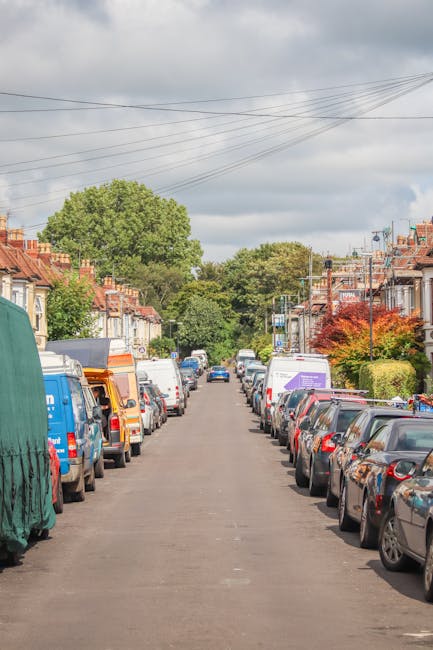 A residential street during daytime with rows of parked cars on both sides, including a variety of hatchbacks, sedans, and commercial vans. In the foreground, a man wearing a high-visibility vest is lifting a large, cardboard box wrapped in bubble wrap onto a trolley, preparing to load it into a white moving van with its back doors open. The van is positioned on the pavement in front of a driveway, with the street extending into the distance beneath a cloudy sky. The houses on either side are typical terraced properties with brick facades, small front gardens, and some have colorful foliage or trees visible. Overhead, power lines stretch across the scene, and soft natural light illuminates the environment, highlighting the moving process related to house relocations and furniture transport. The scene is captured as part of a professional removal service, referenced by Man with Van Teddington, demonstrating packing, loading, and logistical arrangements for home relocations.