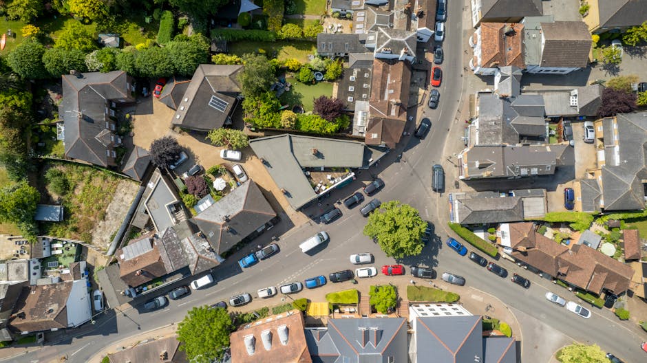 An aerial view of a narrow residential street in Teddington during daylight, showing tightly packed houses with mixed roofing styles, including tiled and flat roofs, lining the street. The street is partially blocked by a line of parked cars, with some vehicles moving or waiting to load or unload. Several houses have small front gardens or yards, with visible trees, shrubs, and garden furniture. A delivery van is parked on the street, and a few residents are seen carrying boxes or furniture towards their homes, reflecting a home relocation or moving process. Man with Van Teddington's company vehicle is also visible, implying professional removals or furniture transport activity. The scene captures the logistics of packing and loading items, highlighting the challenges of navigating narrow roads during house moves in the area.