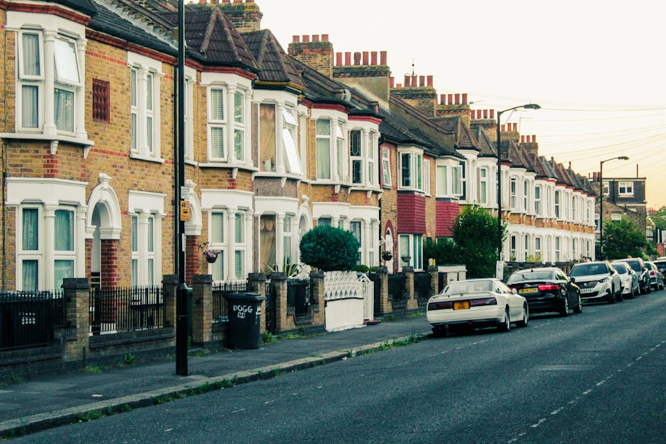 A busy street scene in Teddington with a row of parked cars along the curb on the left side, adjacent to a line of multi-storey buildings with storefronts and awnings, some of which have signage visib