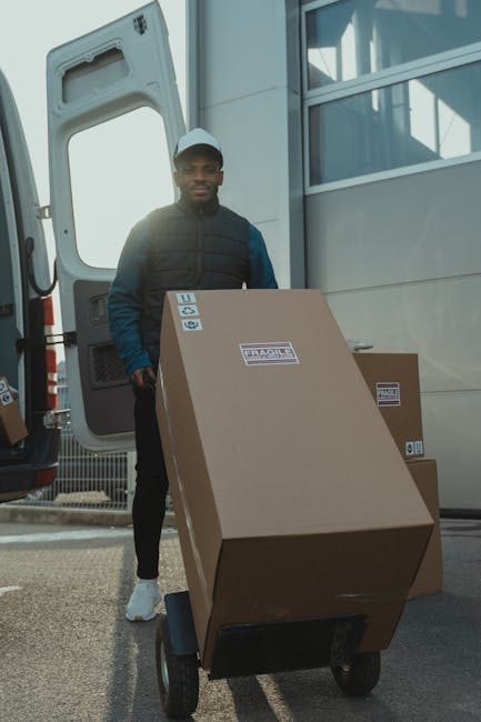 A man wearing a dark jacket, blue sleeves, and a white cap stands outdoors beside a large white van with an open rear door. He is holding a hand truck loaded with a large cardboard box marked with 'Fragile' labels and handling straps, positioned on a black wheeled platform. The scene captures the loading process during a home relocation or removal service, with additional cardboard boxes visible in the background inside the van. The environment appears to be an outdoor area with natural lighting, and the setting indicates an active furniture transport or packing and moving operation as part of the services offered by Man with Van Teddington, consistent with their focus on house removals and quick, efficient moving solutions.