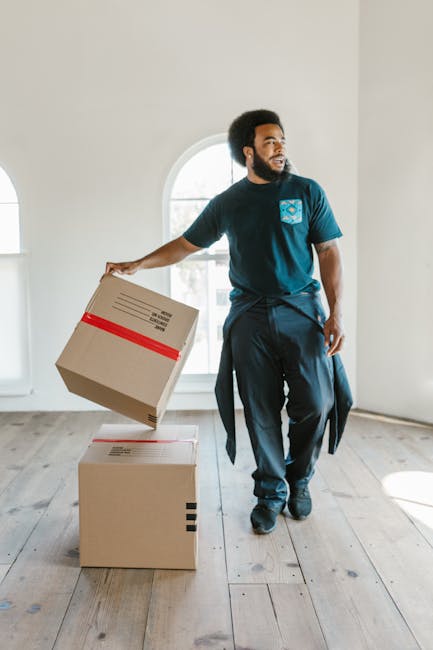 A man with a beard and curly hair, wearing a blue t-shirt with a logo on the chest and dark pants, is standing inside a bright, spacious room with white walls and a large arched window letting in natural light. He is holding a cardboard box with red tape around it in his right hand, preparing to place it on another closed cardboard box on the wooden floor. The room appears to be in the process of a home relocation, with minimal furniture visible. The scene captures the packing and moving process, likely linked to professional removal services by Man with Van Teddington, focused on efficient and careful furniture transport within a house or estate. The environment is well-lit, and the background shows the interior space awaiting further packing or loading activities as part of an urgent or same-day move service in Teddington.